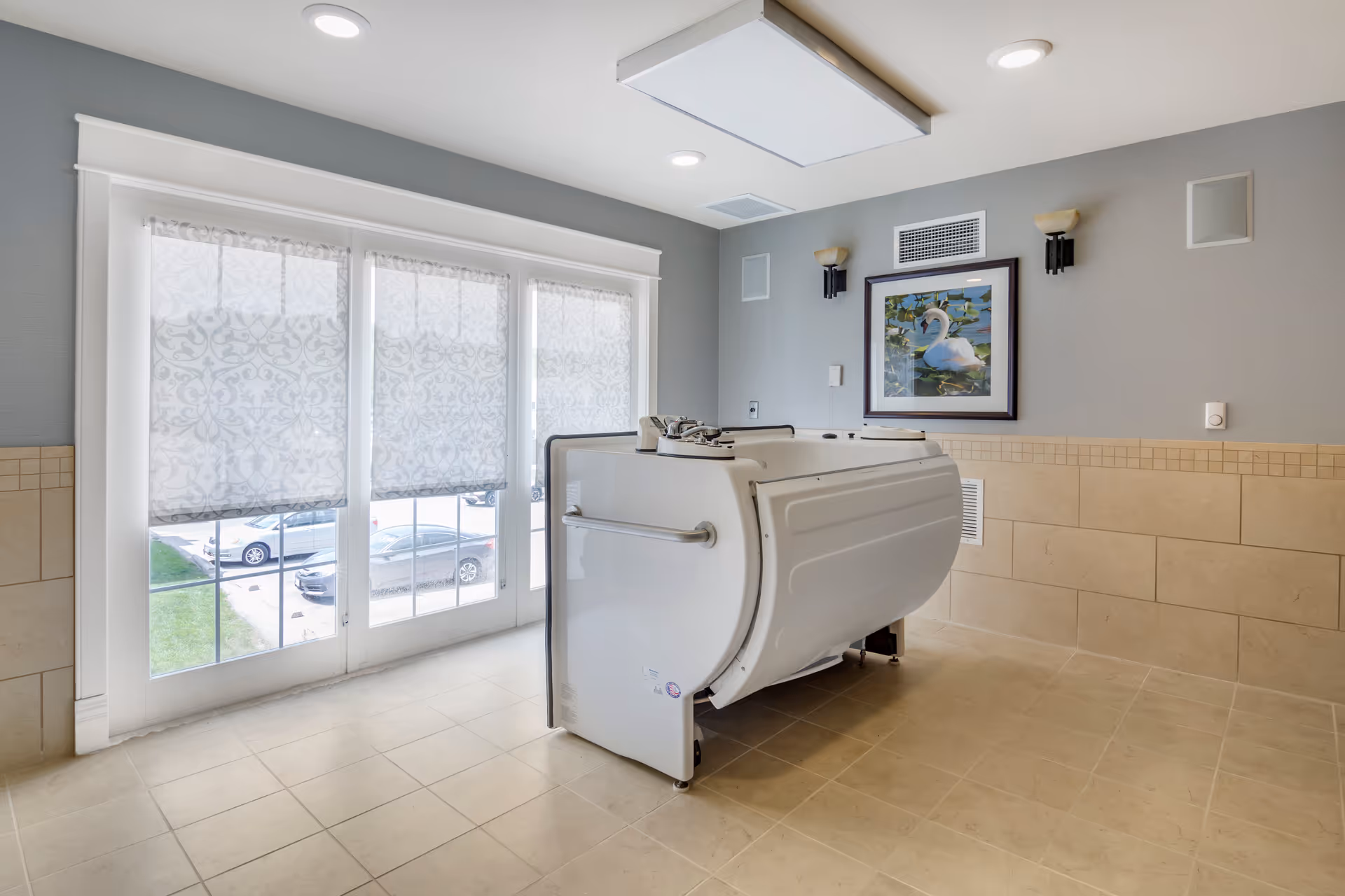 A clean, well-lit room with beige tiled floors and walls, featuring a white walk-in bathtub in the center. The room has large windows with patterned blinds partially drawn, allowing natural light to enter. On the wall above the bathtub, there is a framed picture of a swan on water, flanked by two wall-mounted light fixtures.