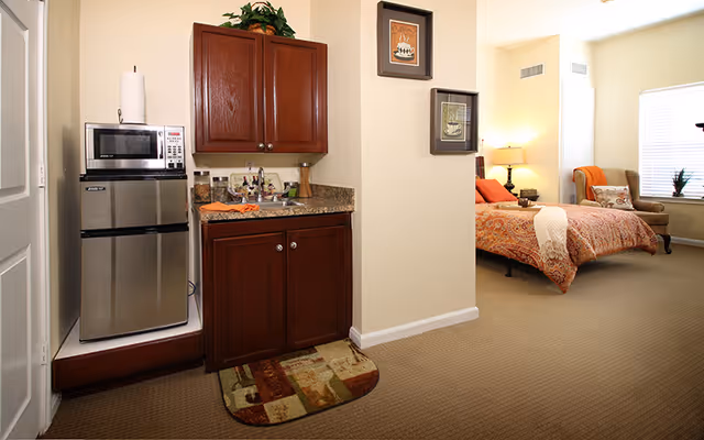 Studio apartment interior with a kitchenette (microwave, mini fridge, sink) and a bed with an armchair in the background.
