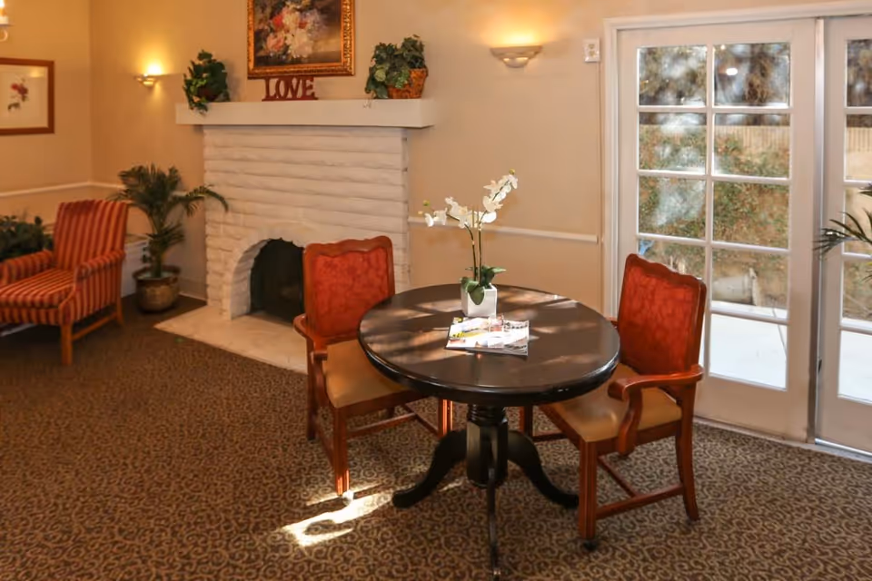 A cozy sitting area in an assisted living facility featuring a round wooden table with a white orchid centerpiece and magazines on top. Two red upholstered chairs with wooden arms are positioned around the table. In the background, there is a white brick fireplace with a decorative 'LOVE' sign and plants on the mantel. To the right, French doors lead outside, allowing natural light to fill the room. The carpet has a patterned design, and the walls are painted a warm beige with framed artwork and wall sconces providing soft lighting.