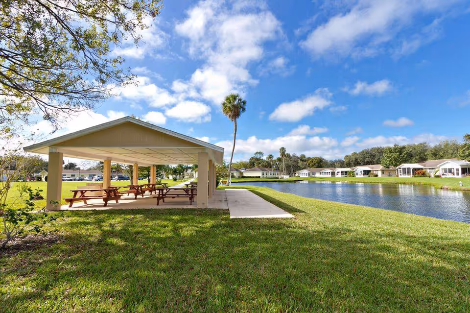 Outdoor covered picnic area with several wooden picnic tables on a concrete slab next to a pond, surrounded by green grass and trees under a partly cloudy blue sky. Residential buildings are visible in the background.