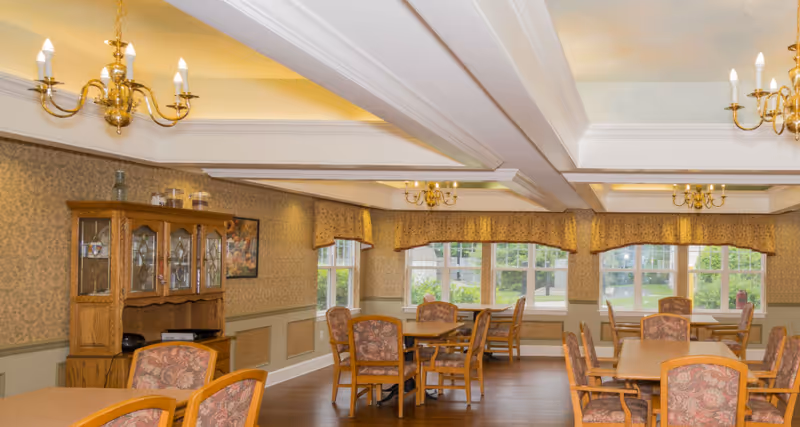 Traditional dining room with wooden tables, upholstered chairs, chandeliers, and windows overlooking greenery.
