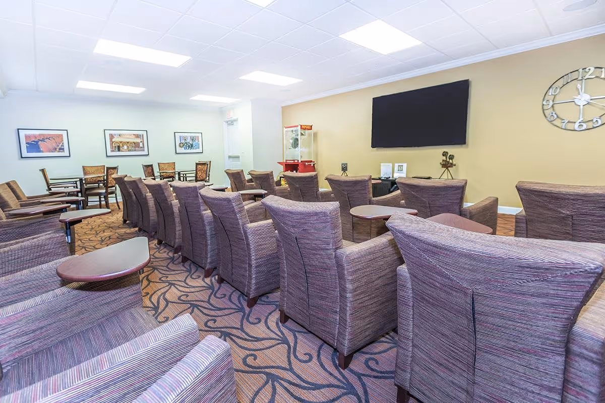 A spacious common area with multiple rows of comfortable upholstered chairs facing a large flat-screen TV mounted on a beige wall. The room has a patterned carpet, a popcorn machine in the corner, a large wall clock, and framed pictures on the opposite wall. There are also several small tables between the chairs.