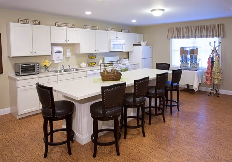 Bright communal kitchen with a large island surrounded by five barstools, white cabinets, appliances, and a beverage station by the window.