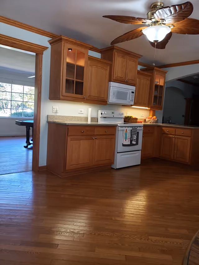 A kitchen with wooden cabinets, a white stove with oven, a white microwave above the stove, and granite countertops. There is a ceiling fan with leaf-shaped blades and a light fixture. The kitchen has hardwood floors and an open doorway leading to a room with a large window and a pool table.