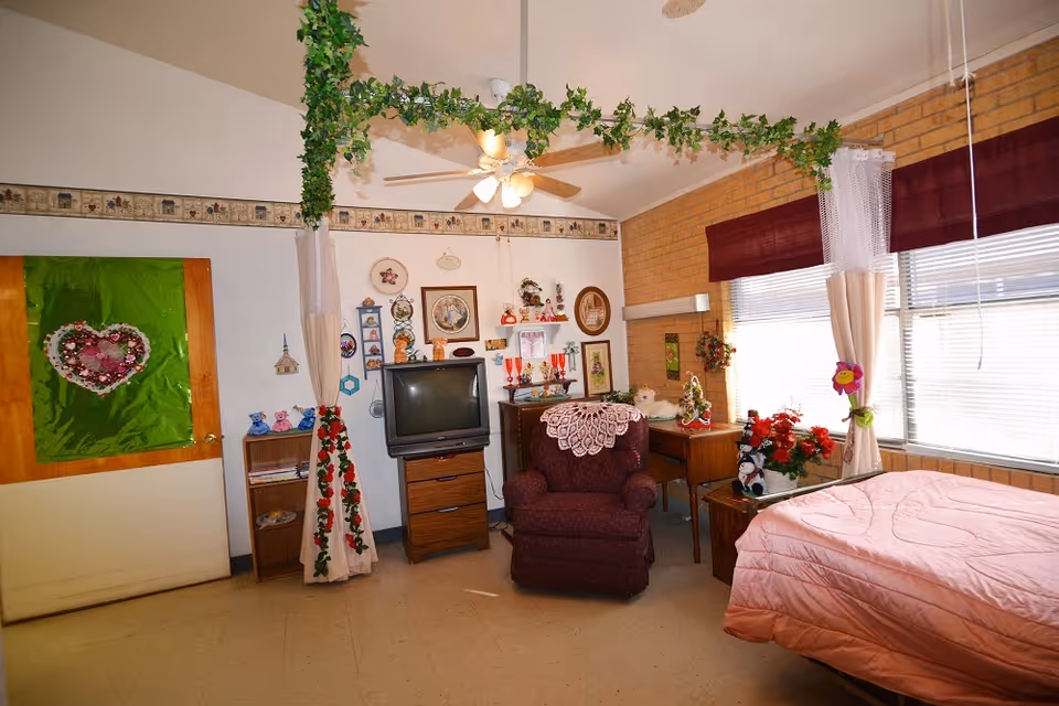 A cozy, decorated bedroom in a nursing and rehab facility featuring a bed with a pink quilt, a maroon armchair with a white doily on the back, a wooden dresser with an older-style TV, a small desk with holiday decorations, and windows with maroon valances and white curtains. Green leafy garlands hang from the ceiling, and the walls are adorned with various framed pictures and decorative items.