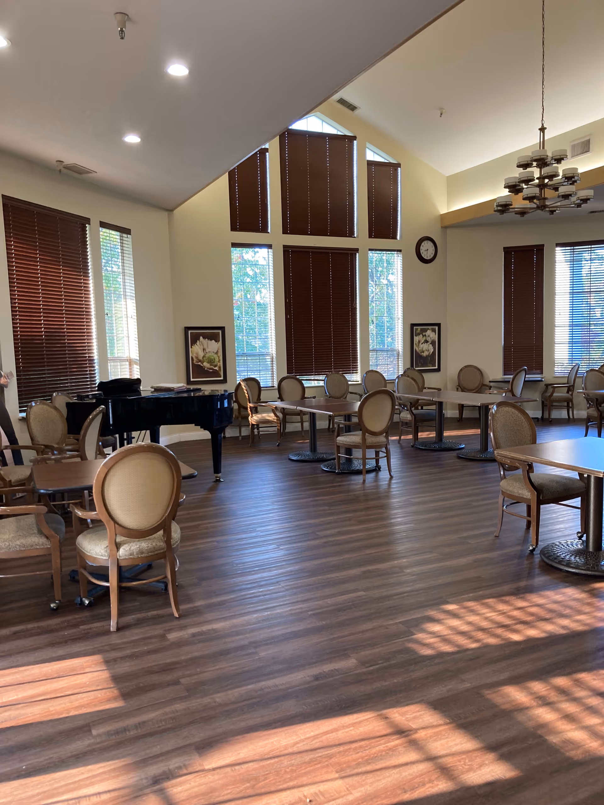 Spacious dining/activity room with round tables and upholstered chairs, a grand piano, tall windows with blinds, and a chandelier.
