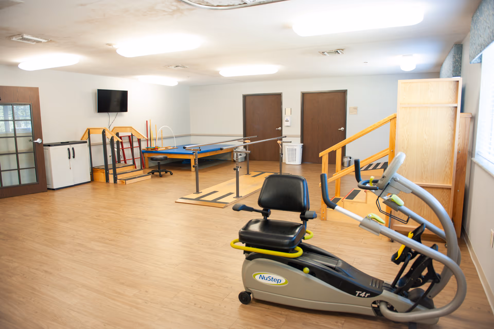 A rehabilitation room with exercise equipment including a NuStep T4r recumbent cross trainer, parallel bars for walking practice, a small set of stairs with a handrail, and a blue therapy table. The room has wooden flooring, white walls, two closed brown doors, a wall-mounted TV, and a window with blinds letting in natural light.