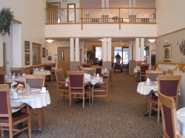 A spacious dining room in a senior living facility with multiple round tables covered with white tablecloths, each set with cups, plates, and utensils. Wooden chairs with purple cushions surround the tables. The room features a high ceiling with a balcony railing above, beige walls, and carpeted floors. There are framed pictures on the walls and some floral decorations on the tables.