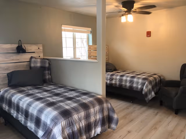 Two single beds with gray plaid bedding in a shared assisted-living bedroom separated by a half wall, with a window, ceiling fan, and an armchair.