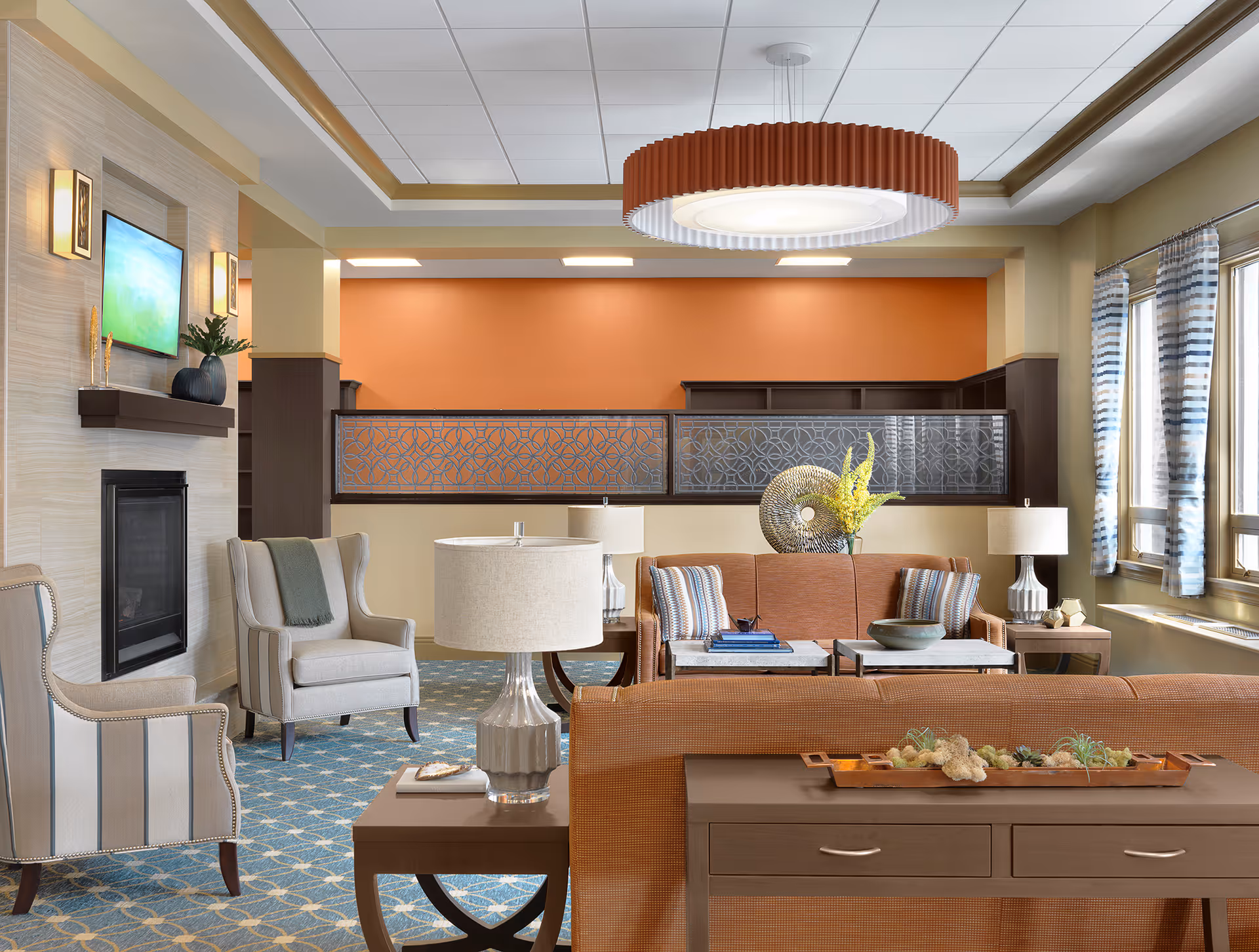 Bright furnished common lounge with sofas, armchairs, side tables, lamps and large windows in a senior living community.