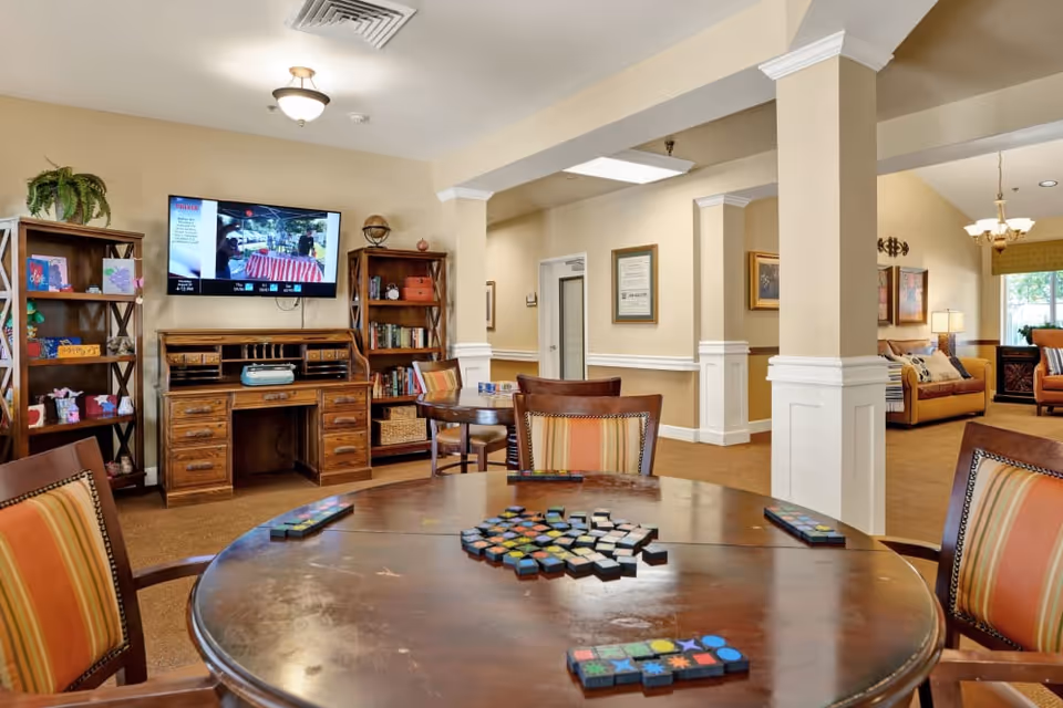 A cozy common area in a senior living facility featuring a round wooden table with a tile game set up, surrounded by chairs with striped cushions. In the background, there is a wooden desk with shelves on either side filled with books and decorative items. A flat-screen TV is mounted on the wall above the desk. The room has beige walls, white trim, and warm lighting, with a seating area visible further back.