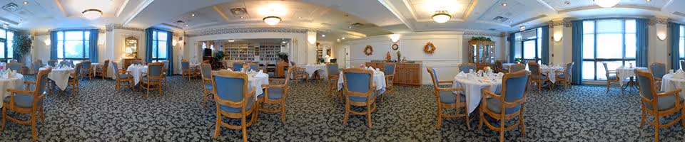 A spacious dining room with multiple round tables covered with white tablecloths, each set with napkins and tableware. The room features blue cushioned wooden chairs, large windows allowing natural light, decorative wreaths on the walls, and a carpeted floor with a patterned design. There are bookshelves and cabinets along the walls, and ceiling lights provide additional illumination.