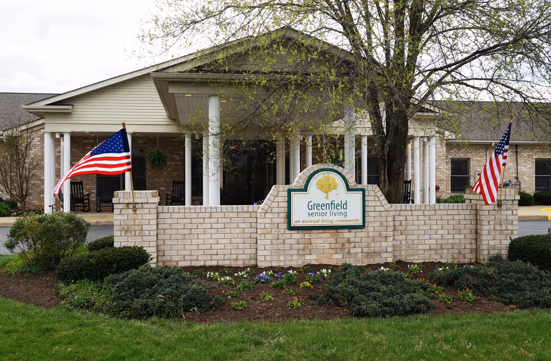 Front exterior view of a senior living facility with a brick sign that reads 'Greenfield senior living an assisted living community'. The building has a covered entrance with white columns, two American flags on either side of the sign, and landscaping with bushes and flowers in front.
