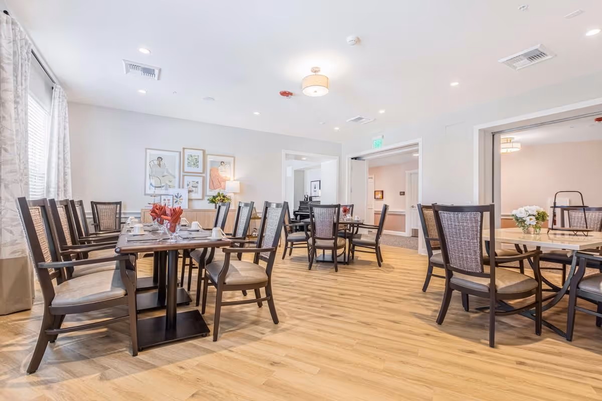 Bright dining room with multiple wooden tables and chairs set for meals in a senior living facility.