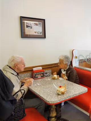 An elderly man and woman sitting across from each other at a small table with red cushioned bench seating in a dining area. They appear to be engaged in conversation while having dessert. The table has a couple of dessert dishes and a small menu or promotional item. A framed picture hangs on the wall behind them.