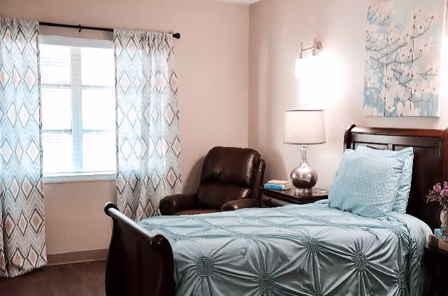 A cozy senior living bedroom featuring a single bed with a light blue patterned bedspread and matching pillow. Next to the bed is a wooden nightstand with a silver lamp and some books. A comfortable dark brown leather armchair is positioned near a window with patterned curtains. A floral painting hangs on the wall above the bed.
