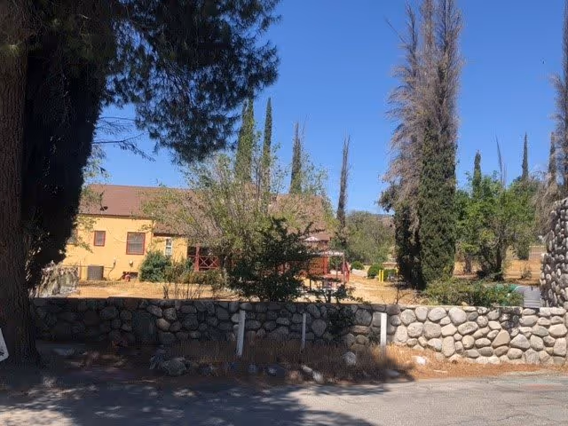 Stone wall and driveway in front of a yellow two-story building with trees, a red porch, and a clear blue sky.