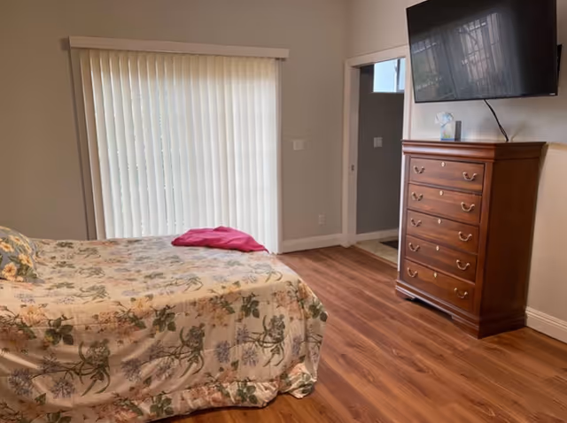 A bedroom with a bed covered in a floral bedspread and a pink pillow. There is a wooden dresser with multiple drawers against the wall, and a flat-screen TV mounted above it. Vertical blinds cover a window or sliding door, and the floor is wood.