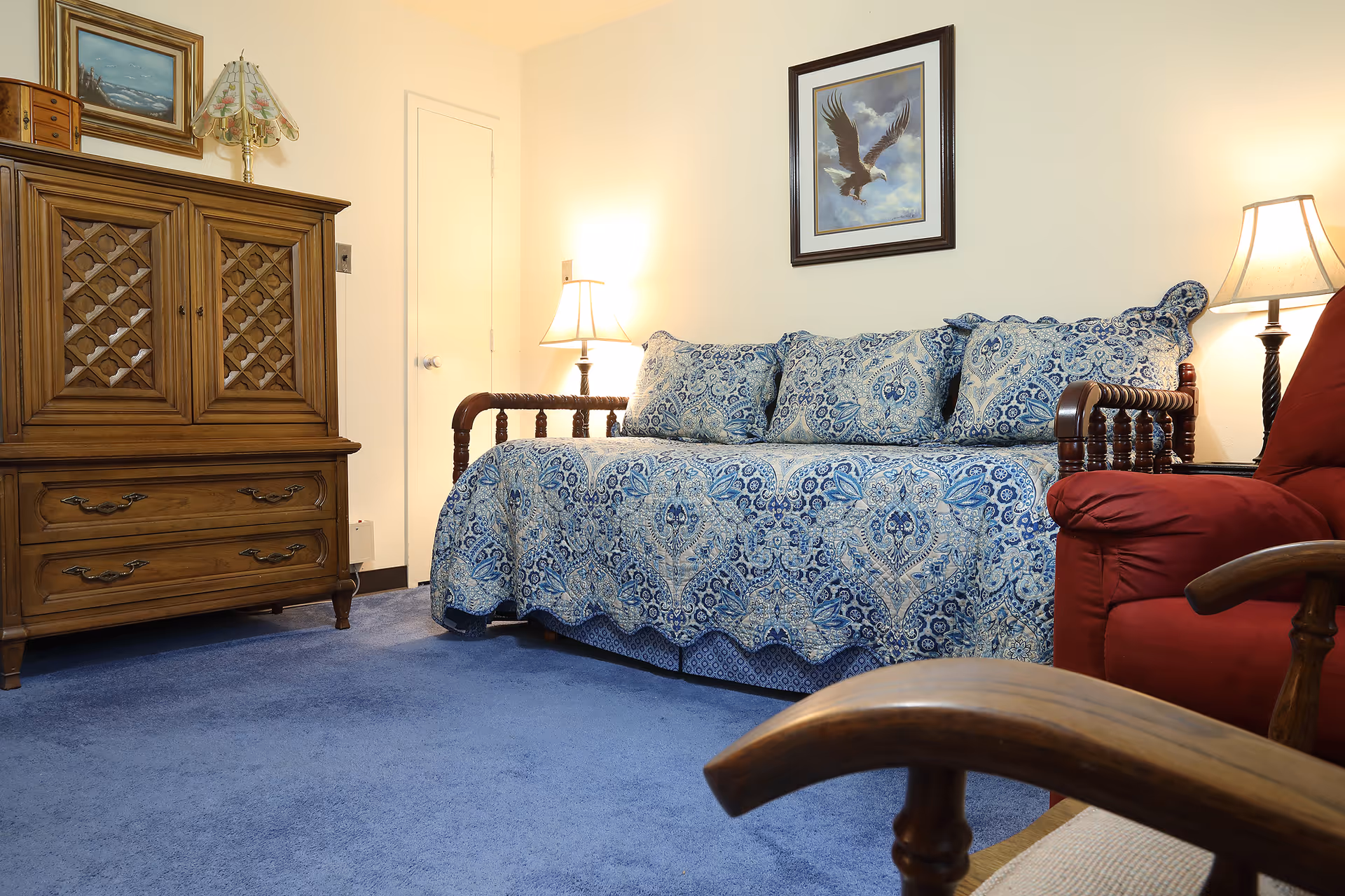 A cozy living room with a blue patterned daybed covered with matching pillows, a wooden cabinet with drawers and lattice doors, a red armchair, two table lamps providing warm light, and a framed picture of an eagle on the wall.