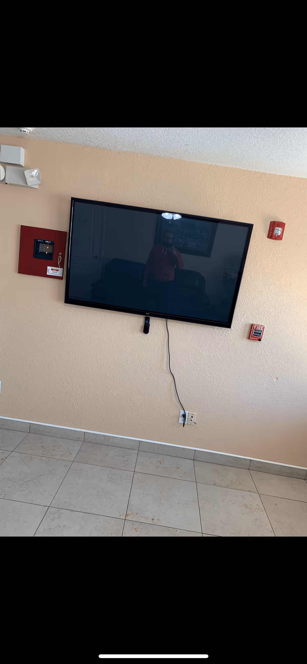 A wall-mounted flat screen TV with a remote control hanging below it. The TV is off and reflects a person taking the photo. The wall is beige with a red fire alarm pull station and a red fire alarm control panel mounted on it. The floor is tiled with light-colored tiles.