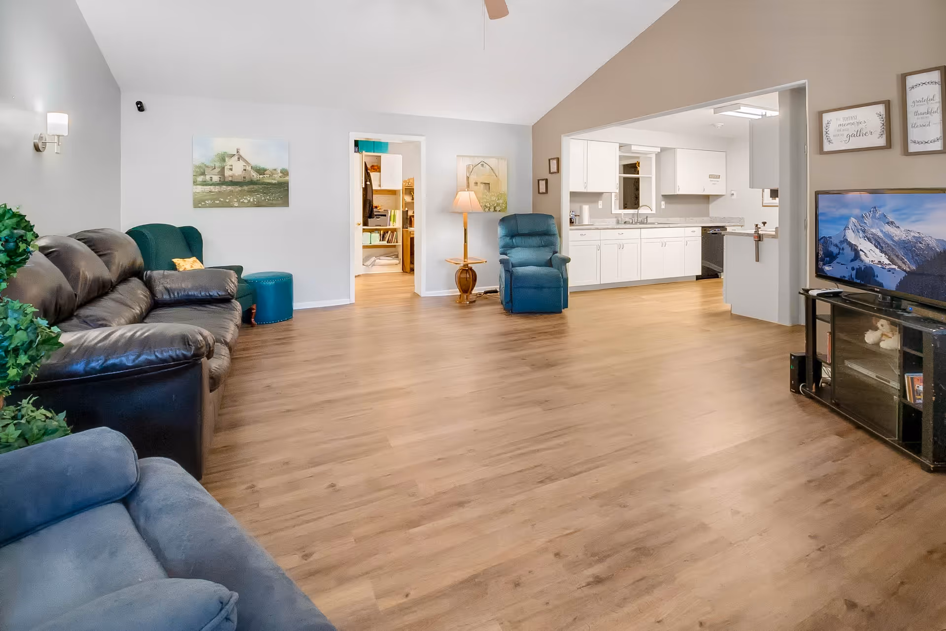 A spacious living room area with wooden flooring, featuring a black leather sofa, a blue recliner chair, a green armchair with a matching ottoman, a floor lamp, and a flat-screen TV on a black stand. The room opens into a white kitchen with cabinets and countertops. The walls are decorated with framed pictures and a ceiling fan is visible.