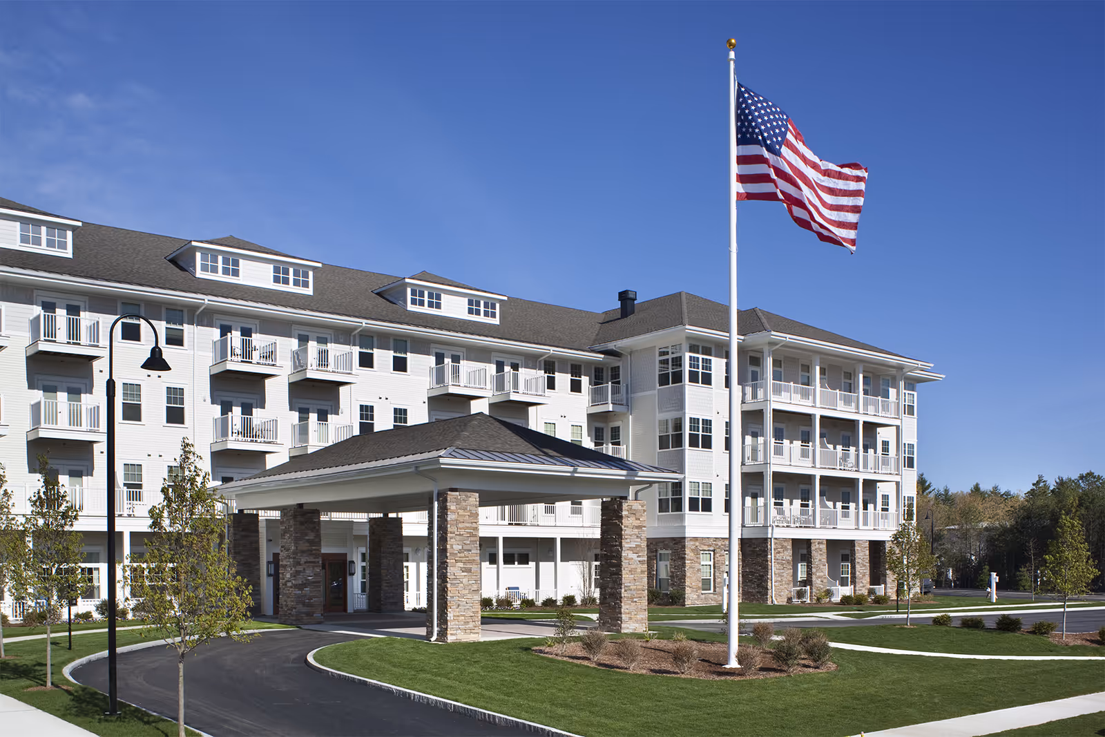 Exterior view of a multi-story senior living facility with white siding and stone accents. The building features multiple balconies and a covered entrance supported by stone pillars. An American flag flies on a flagpole in front of the building, and there is a paved driveway and landscaped lawn with young trees and shrubs under a clear blue sky.