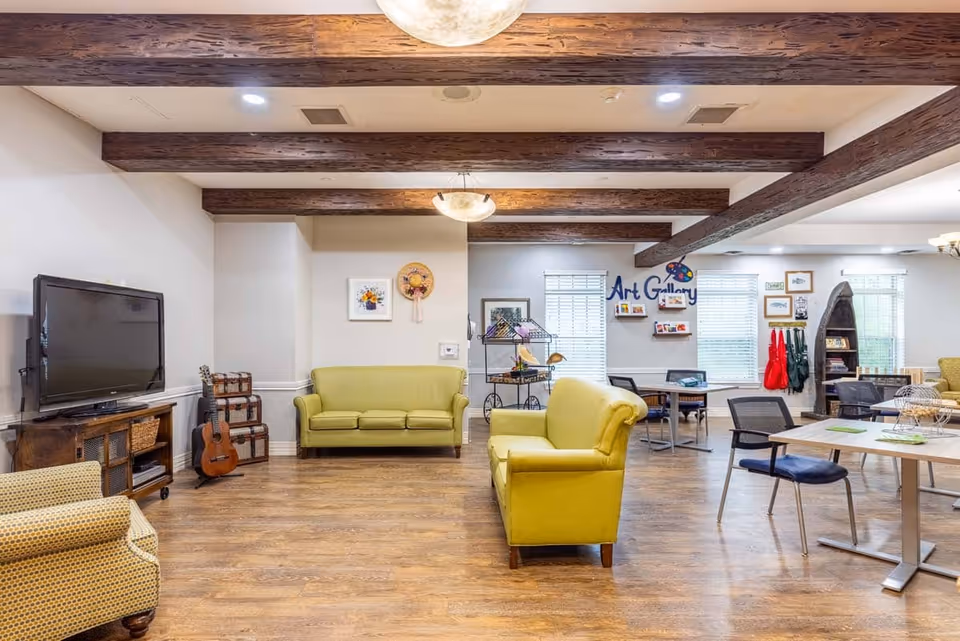 Spacious communal lounge with yellow sofas, a TV, tables and chairs, and an 'Art Gallery' activity area beneath exposed wooden ceiling beams.