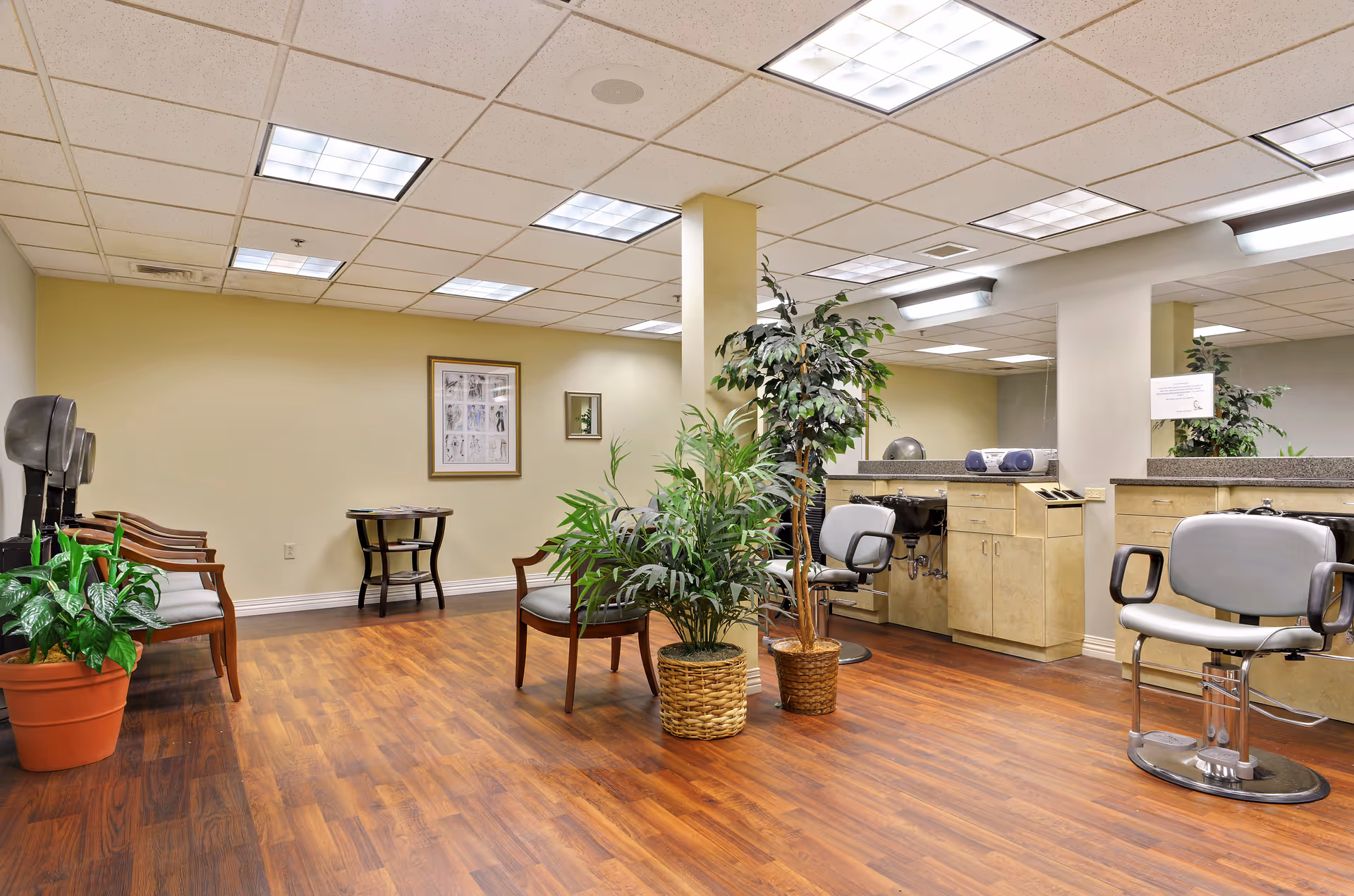 Interior view of a hair salon area in a senior living facility with wooden flooring, salon chairs, hair dryers, plants in pots, a small table with magazines, and large mirrors on the walls.