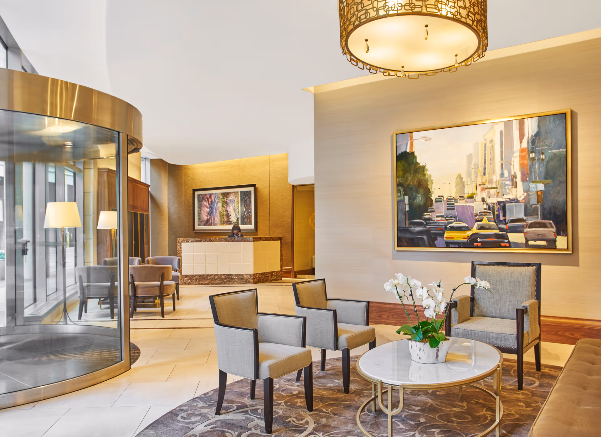 A bright and modern lobby area with a revolving glass door on the left, a reception desk with a person behind it in the background, and a seating area in the foreground featuring four upholstered chairs around a round marble coffee table with a white orchid plant. A large colorful painting of a city street with cars hangs on the wall, and a decorative ceiling light fixture is visible above.