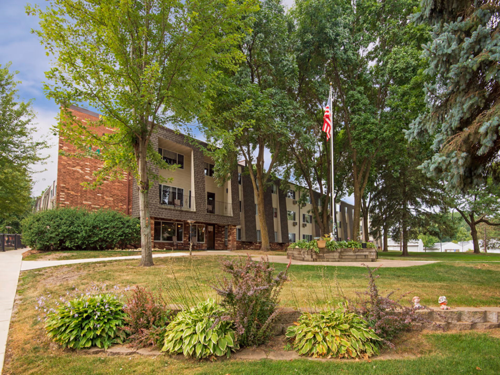 Exterior view of a three-story senior living facility building named Kilkenny Court, surrounded by large trees, green grass, and landscaped bushes. An American flag is displayed on a flagpole in front of the building.