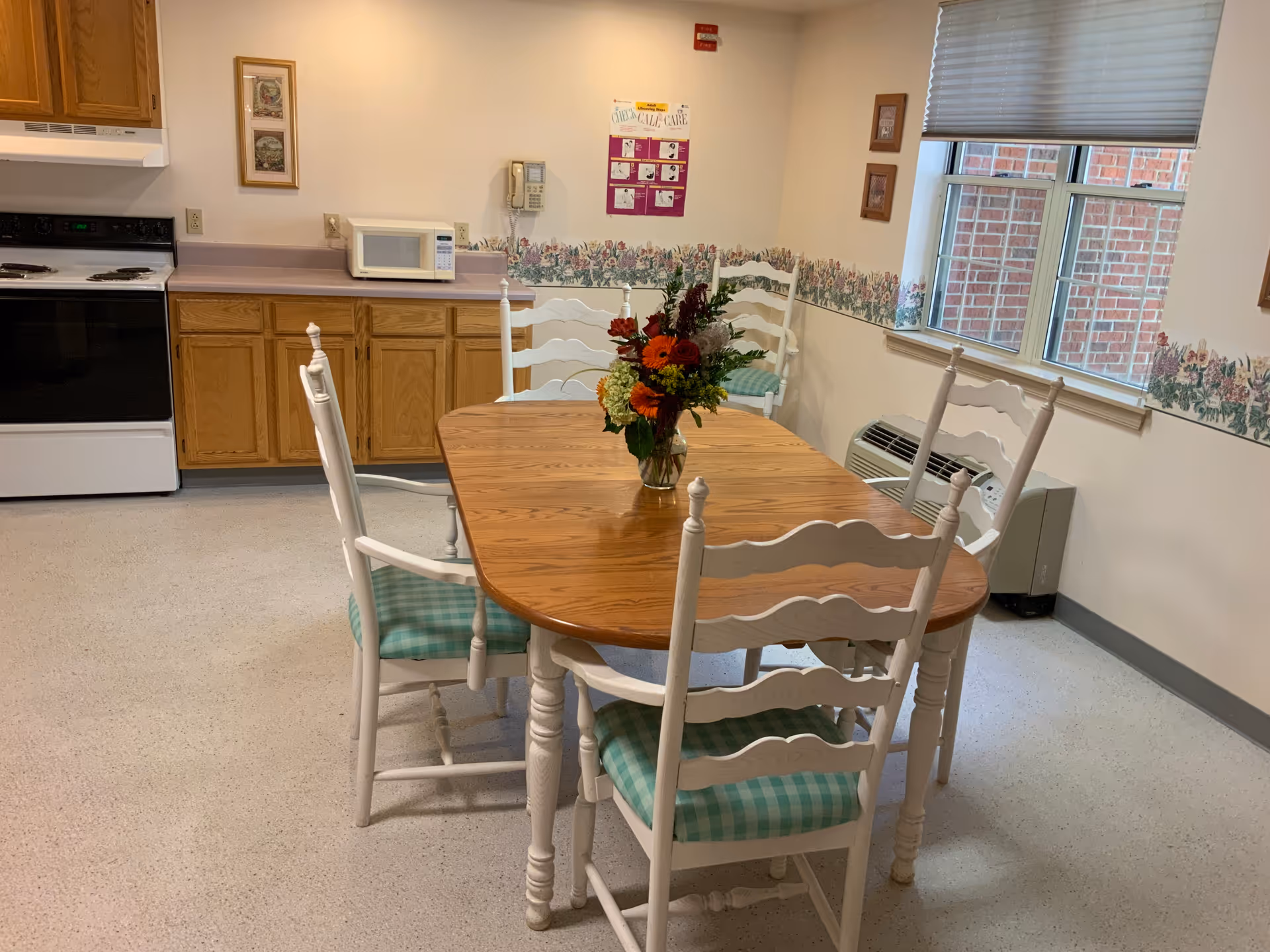 Communal kitchen-dining area with a wooden table and six white chairs with green cushions and a vase of flowers, cabinets, stove and microwave.