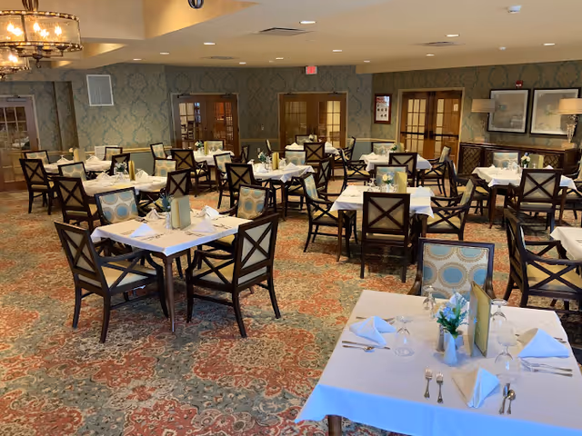 Dining room with numerous neatly set tables and upholstered chairs on a patterned carpet in a senior living community.