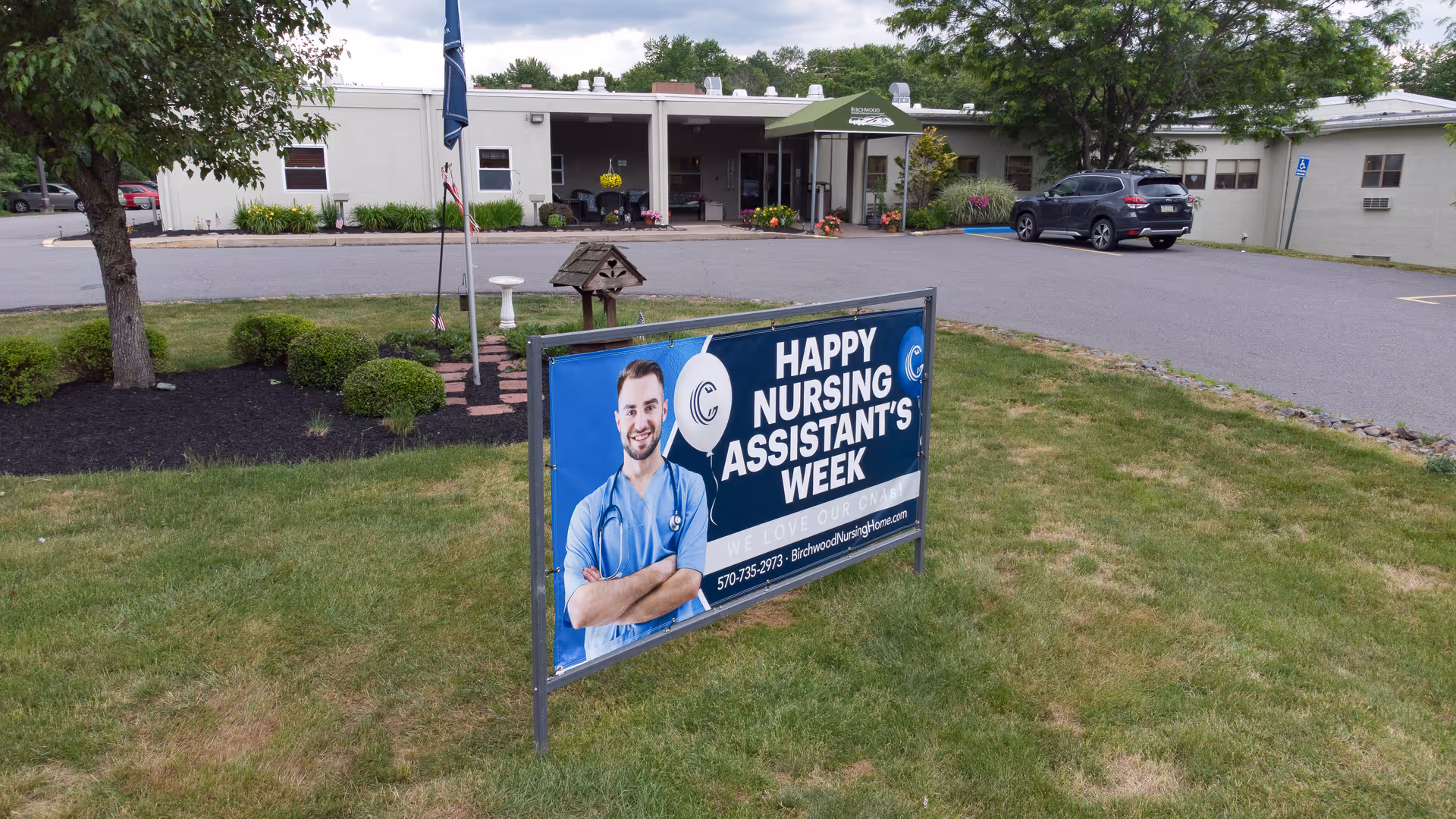 Front exterior of Birchwood Rehabilitation and Healthcare Center with a lawn sign that reads 'Happy Nursing Assistant's Week' and a smiling healthcare worker image.