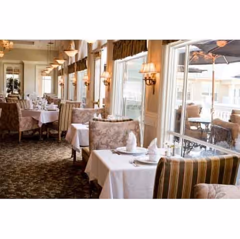 Elegant dining room with tables covered in white tablecloths, set with plates, napkins, and silverware. The room features large windows with natural light, wall-mounted lamps, and upholstered chairs with floral and striped patterns. Outside the windows, there is a patio area with outdoor seating and umbrellas.