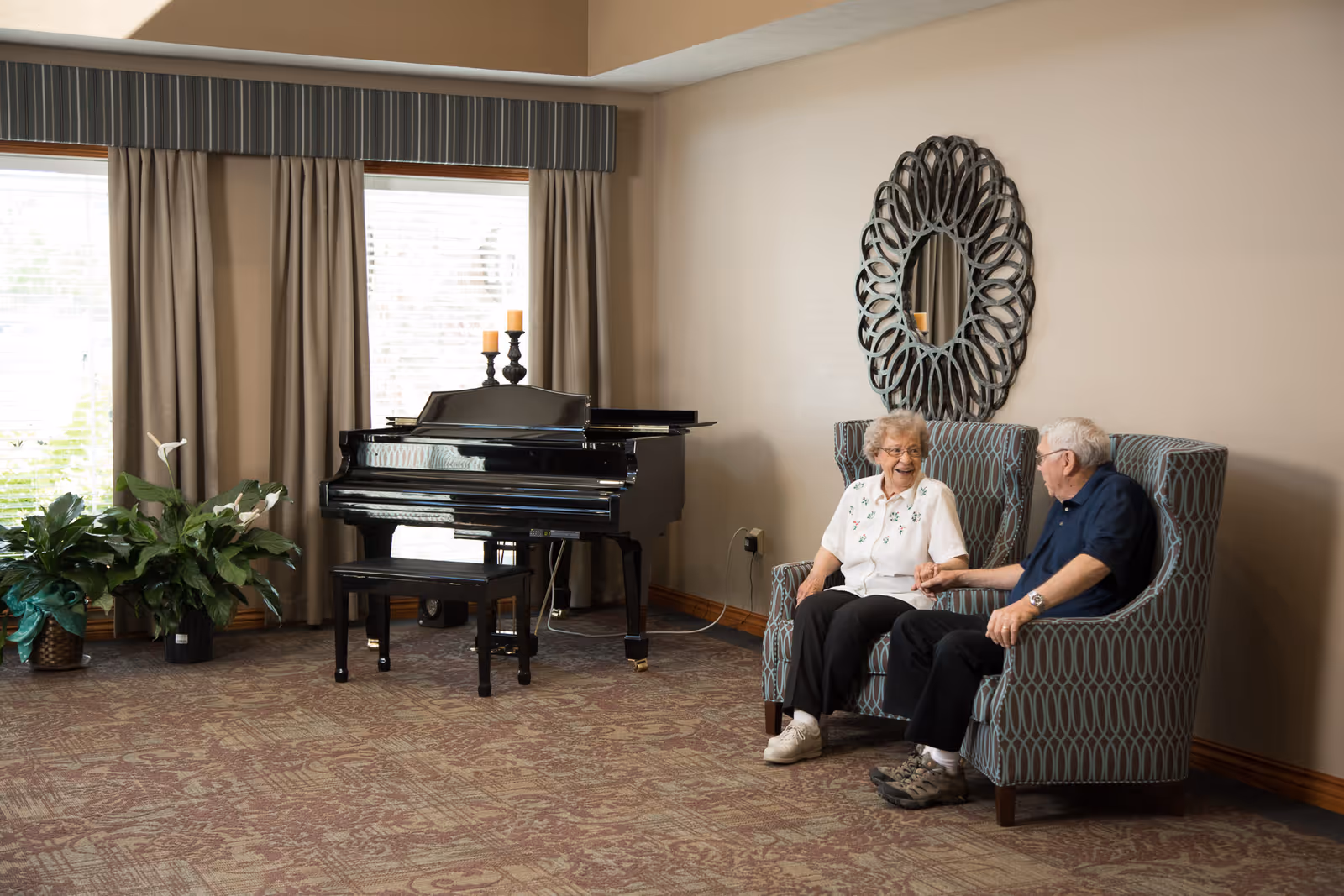 Two elderly residents sit and hold hands on patterned armchairs in a lounge area next to a grand piano and potted plants.