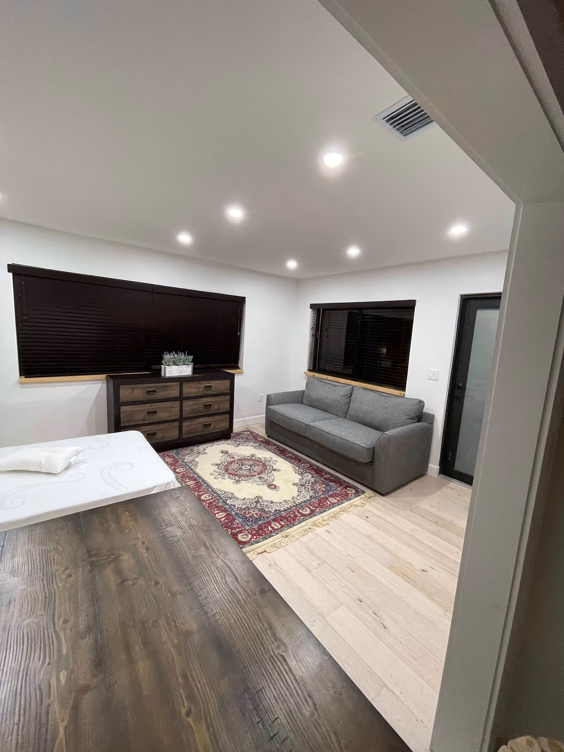 A modern, minimally furnished room with light wood flooring, white walls, and recessed ceiling lights. The room contains a gray sofa, a wooden dresser with six drawers, a patterned area rug, and a wooden table with a white mattress or cushion on it. Two windows with dark blinds and a door with frosted glass are visible.