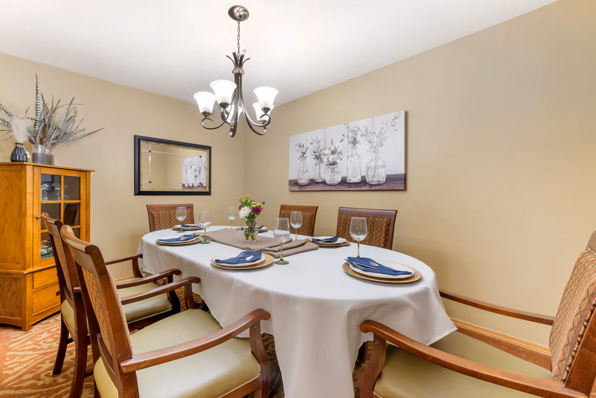 A dining room with an oval table covered with a white tablecloth set for six people. Each place setting has a gold charger plate, a blue napkin, and a wine glass. There is a small flower arrangement in the center of the table. The room has beige walls, a wooden cabinet with decorative vases, a rectangular mirror, and a painting of flowers in glass jars. A chandelier with five lights hangs above the table.