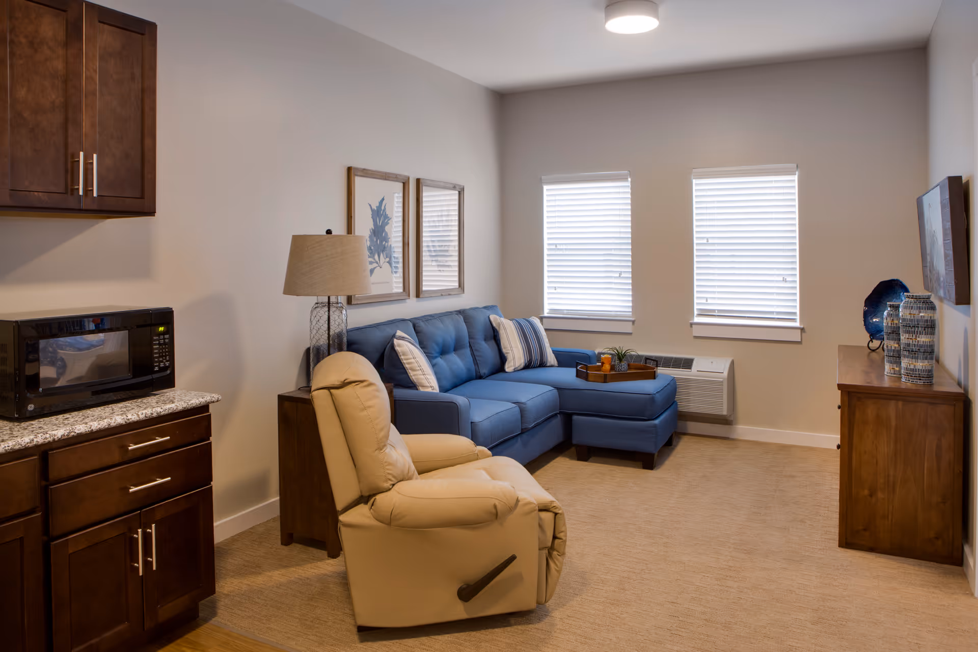 Cozy living room with a blue sectional sofa, beige recliner, wooden cabinets with a microwave, and two windows with blinds.