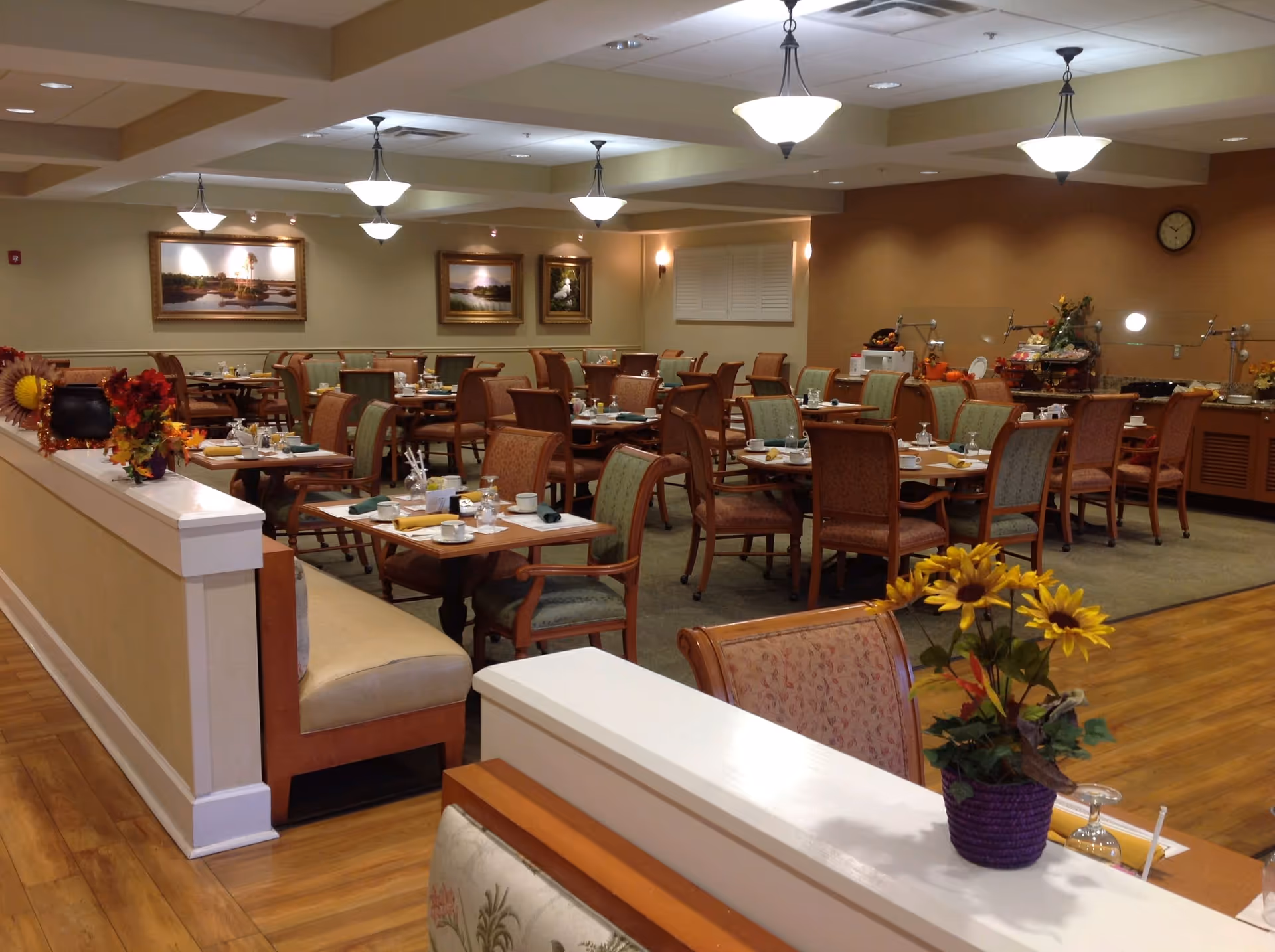 A spacious dining room with multiple wooden tables and chairs arranged neatly. Each table is set with cups, plates, napkins, and utensils. The room is well-lit with hanging ceiling lights and decorated with framed landscape paintings on the walls. There are autumn-themed floral arrangements on some tables and a buffet area in the background with food and serving utensils.