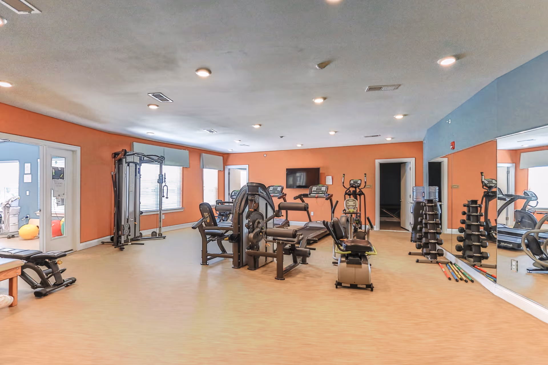 A spacious fitness room with various exercise equipment including treadmills, weight machines, dumbbells, and an elliptical machine. The room has orange and blue walls, large windows with blinds, a wall-mounted TV, and a large mirror on one side. The floor is light-colored, and the ceiling has multiple recessed lights.