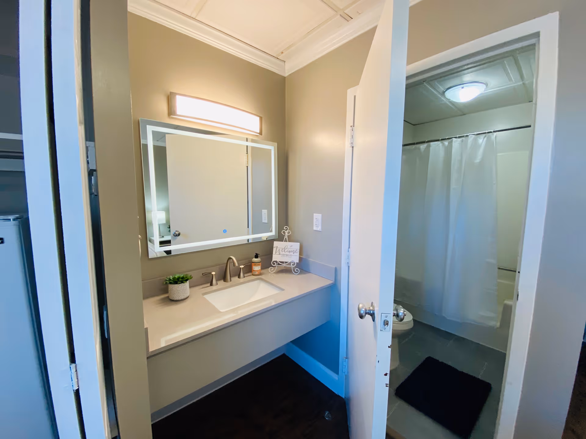 A bathroom vanity with a lighted mirror, sink and faucet, and an open door revealing a toilet and shower with a white curtain.