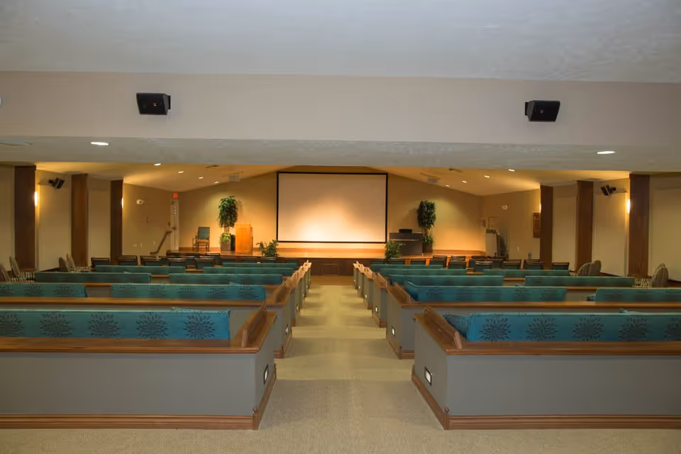 Interior view of a large meeting or presentation room with rows of teal cushioned pew-style seating facing a stage with a large blank projection screen and a wooden podium. The room has beige walls, carpeted floor, and potted plants on the stage.