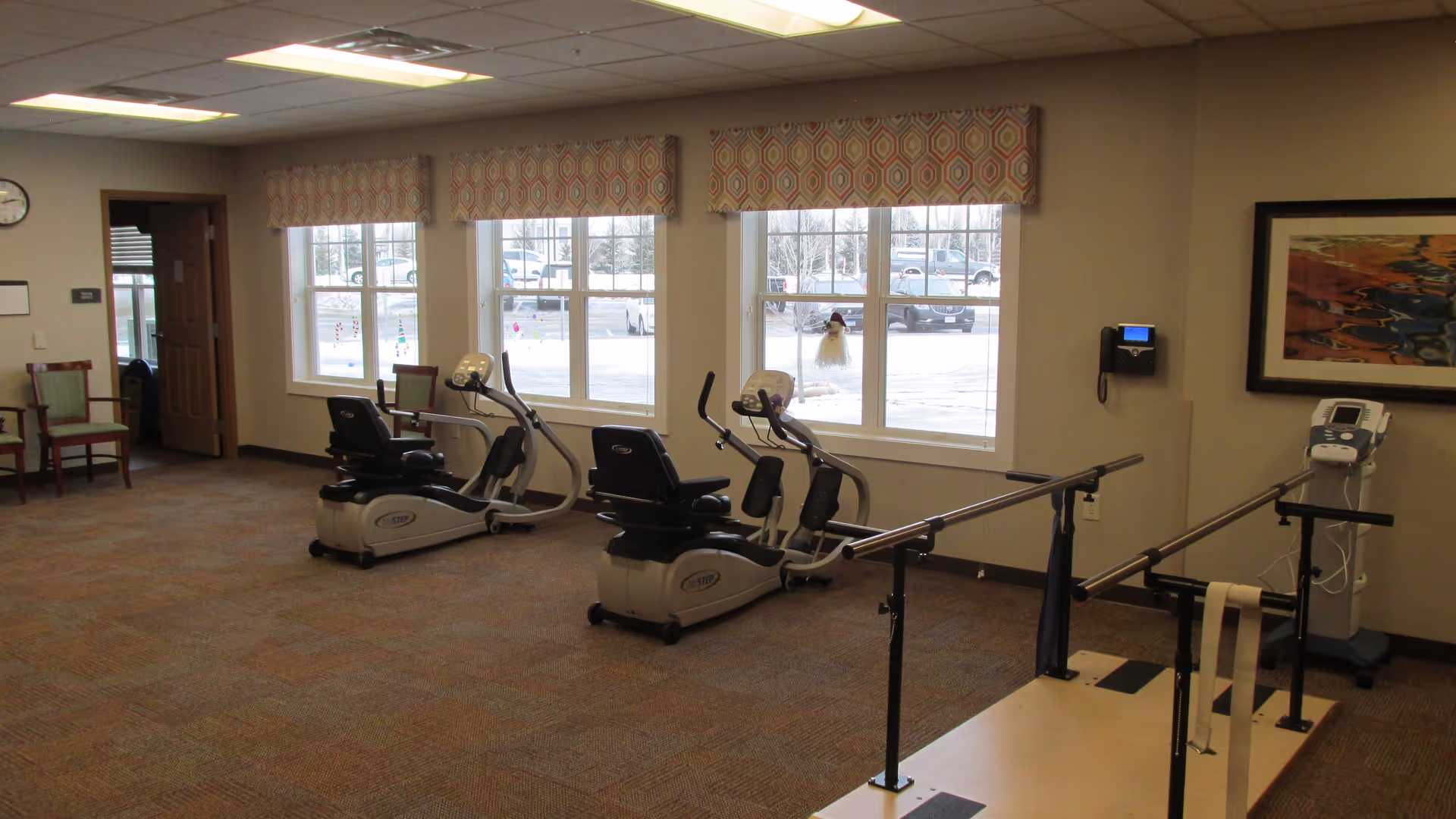 Exercise/therapy room with recumbent stationary bikes and parallel walking bars in front of large windows.