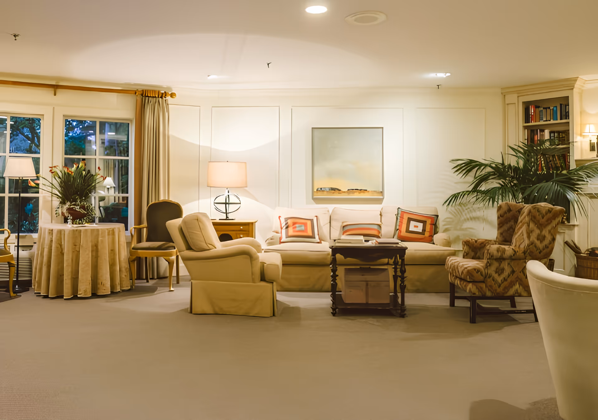 A cozy living room with a beige sofa adorned with colorful square-patterned pillows, two armchairs, a wooden coffee table, a side table with a lamp, a round table with a floral arrangement, large windows with curtains, and built-in bookshelves with plants and books.