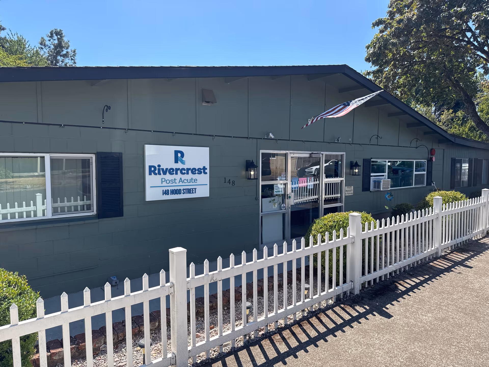 Single-story green building with a white picket fence and a sign reading "Rivercrest Post Acute" by the entrance under an American flag.
