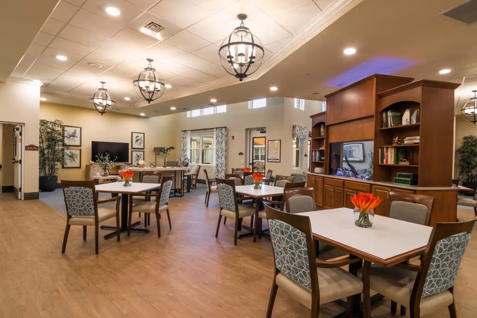 Well-lit communal dining and activity room with multiple tables and chairs, pendant lights, a TV, and wooden shelving.