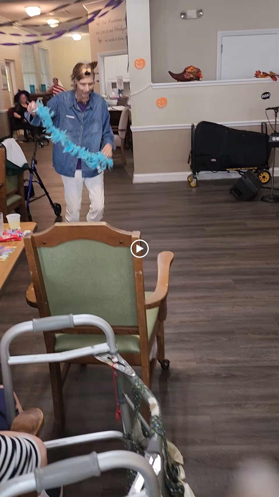 An elderly person wearing a denim jacket and white pants is holding a blue garland in a decorated room with Halloween-themed decorations on the walls. There are chairs, a walker, and other people seated in the background. The room has wooden flooring and light-colored walls.