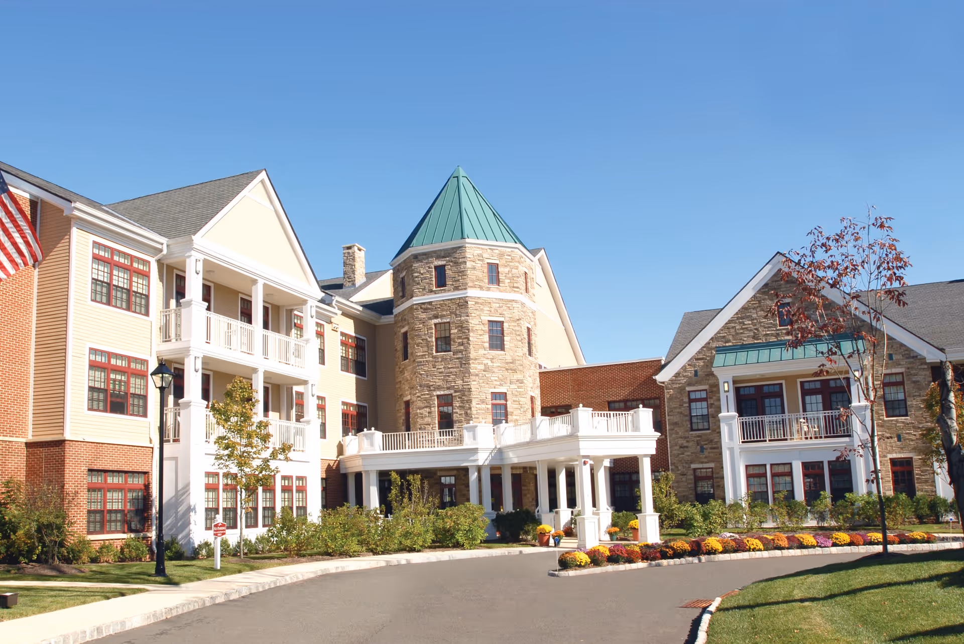 Front entrance of a multi-story senior living building with a stone turret, white balconies, and landscaped driveway under a clear blue sky.