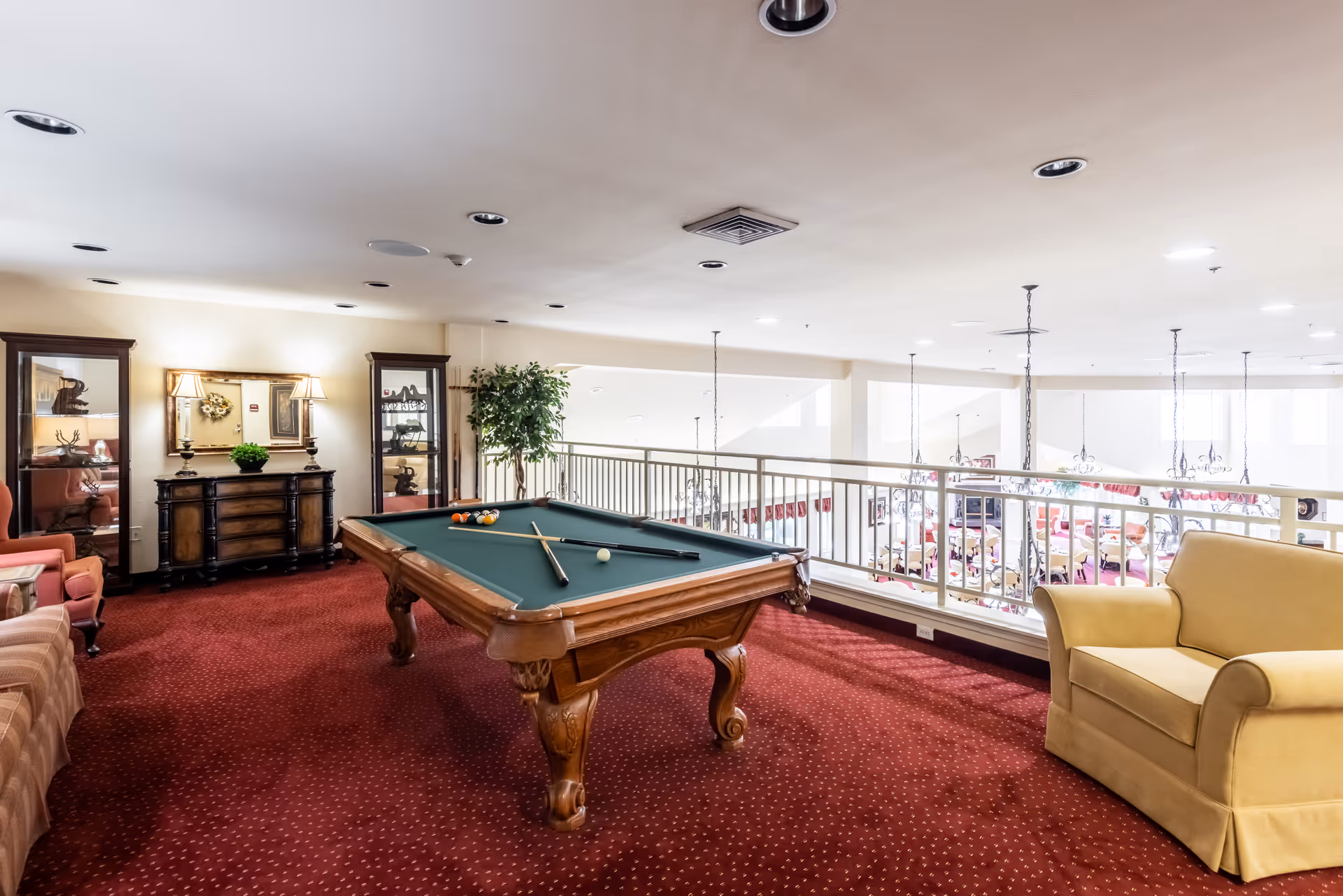 A senior living facility game room with a pool table in the center, two pool cues resting on the table, a yellow armchair on the right, a striped sofa on the left, and two glass display cabinets and a wooden dresser with a lamp and plant against the back wall. The room has red carpet flooring and overlooks a dining area below through a white railing.