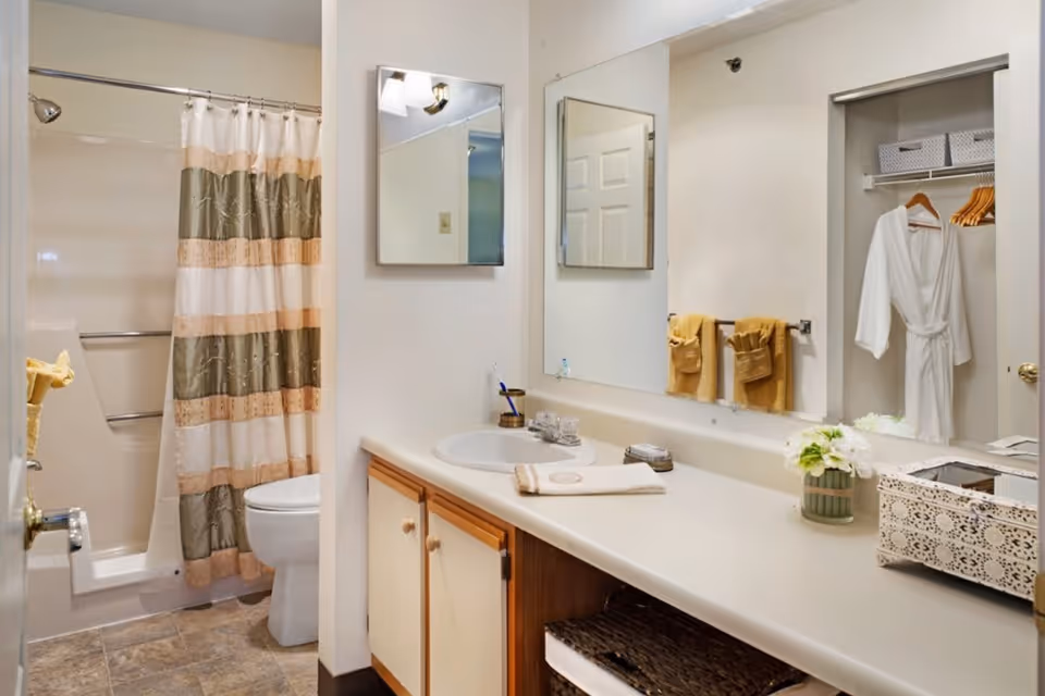 A clean bathroom with a shower and bathtub combination featuring a striped shower curtain in beige, green, and peach colors. There is a toilet next to the shower. The bathroom has a long countertop with a sink, a mirror above it, and a smaller medicine cabinet mirror on the wall. Yellow towels hang on a towel rack, and a white bathrobe is hanging in an open closet. The countertop also has a decorative flower vase and a white lace-patterned box.